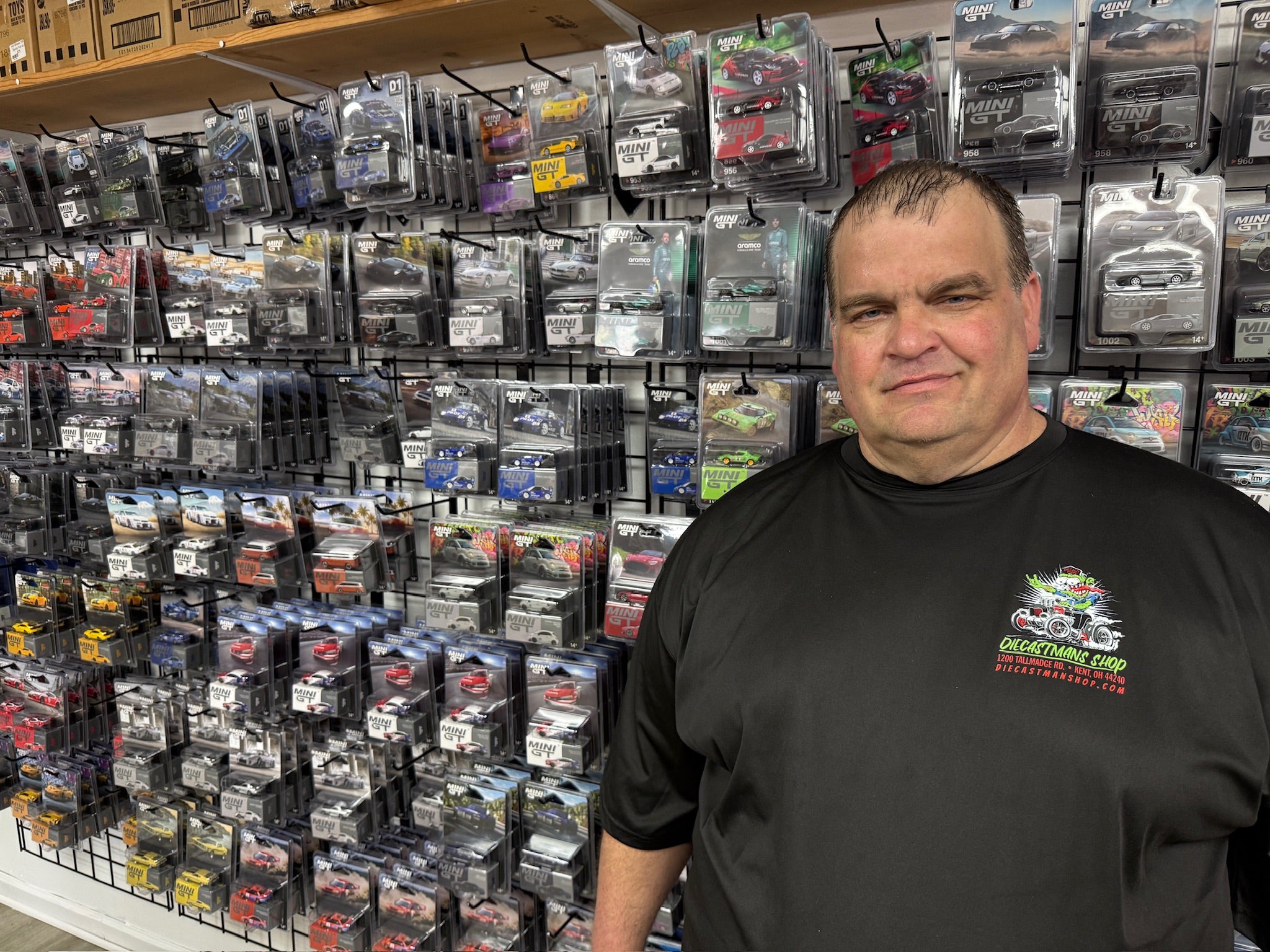 A man wearing a black t-shirt with a 'Diecastman's Shop' logo stands in front of a wall densely packed with packaged die-cast model cars in a retail store.